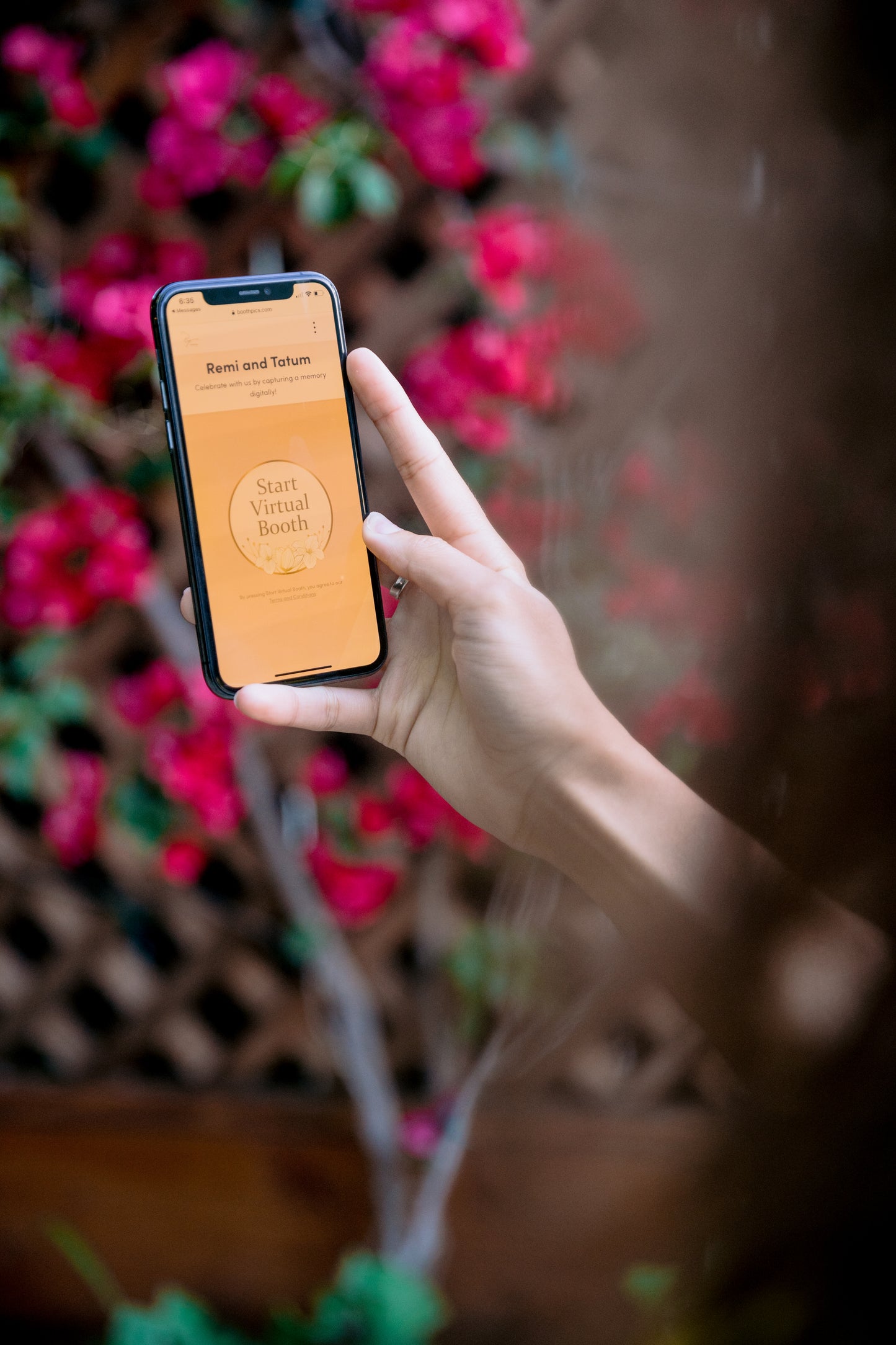 Close-up of a person holding a phone displaying the 'Start Virtual Booth' screen surrounded by flowers.