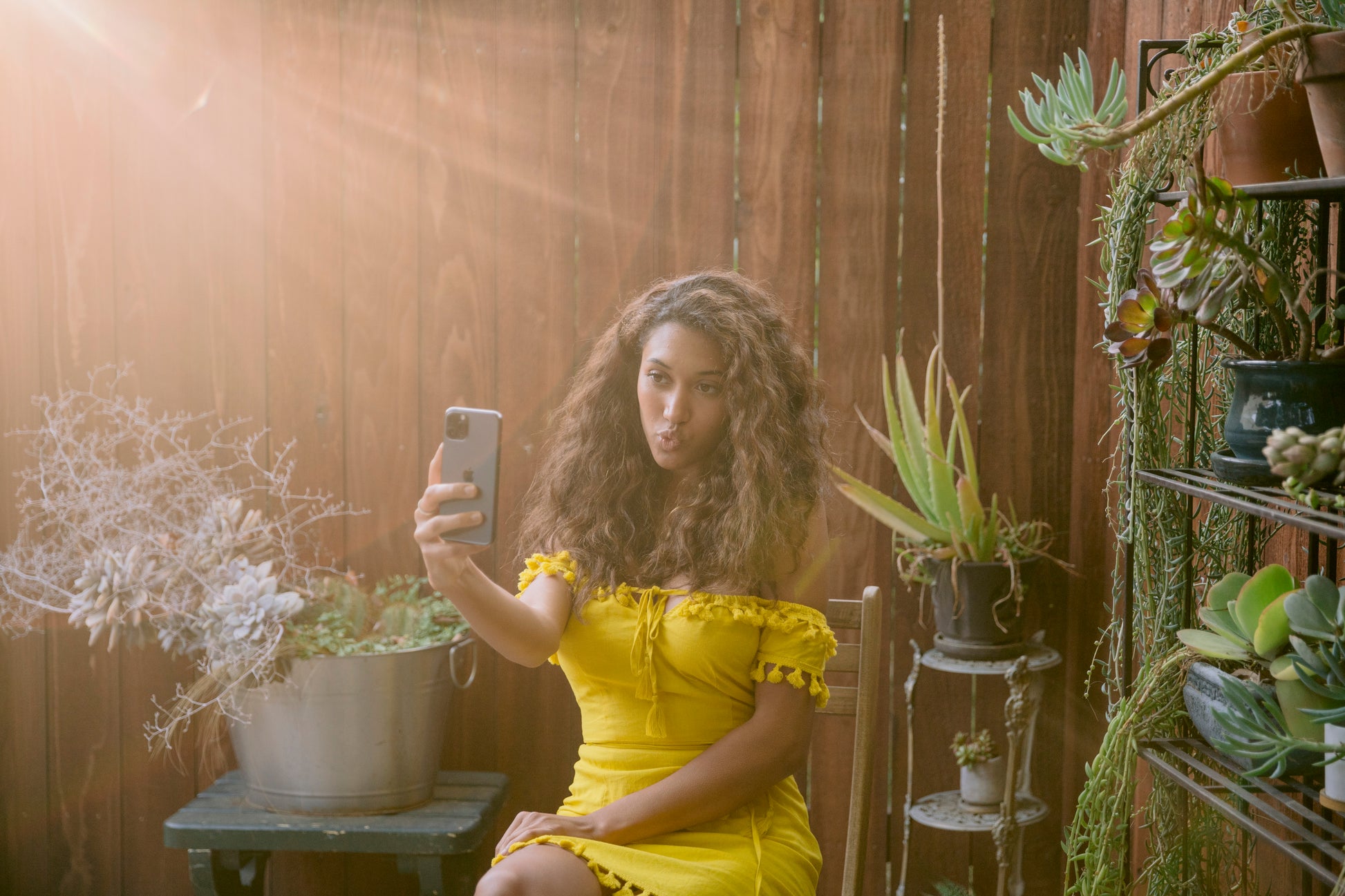 A person in a yellow dress taking a selfie in a sunlit garden setting with green plants.