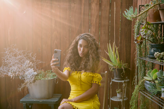 A person in a yellow dress taking a selfie in a sunlit garden setting with green plants.